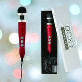 A Doxy Die Cast 3 Wand Massager in Candy Red is featured next to its packaging. The polished device, highlighting its buttons and coiled cable, stands out brilliantly against a background of softly blurred colorful lights.